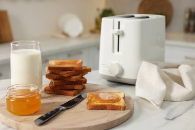 Kahvaltı mutfakta servis edilir. Tost makinesi, çıtır ekmek, bal ve süt beyaz masada.