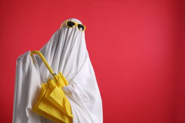 Hayalet kostümlü ve güneş gözlüklü biri kırmızı arka planda sarı şemsiye tutuyor, mesaj için yer var.