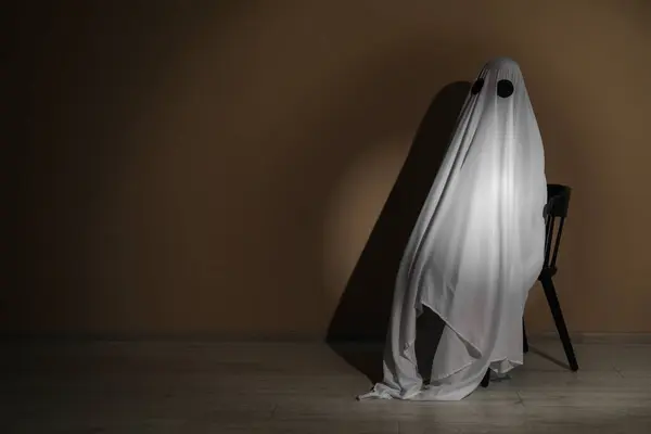 Creepy ghost. Woman covered with sheet sitting on chair near brown wall, space for text