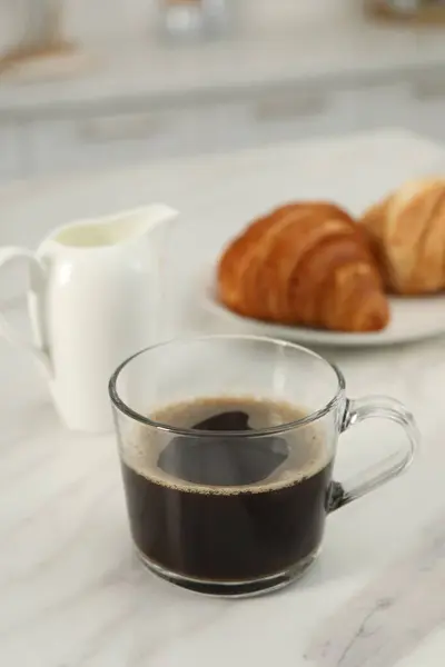 Aromatic coffee in glass cup, pitcher and fresh croissants on white marble table