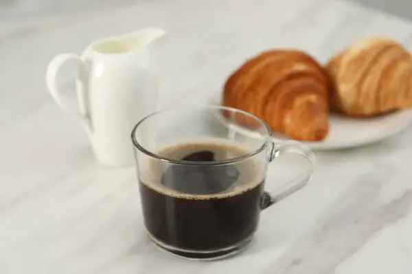 Aromatic coffee in glass cup, pitcher and fresh croissants on white marble table, closeup