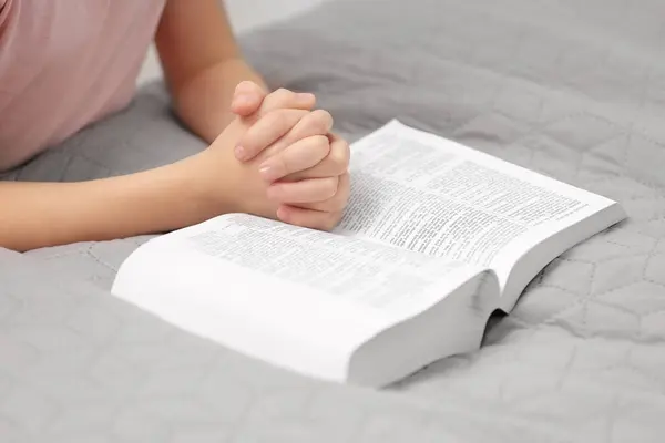 Girl holding hands clasped while praying over Bible indoors, closeup