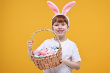Paskalya kutlaması. Tavşan kulaklı ve turuncu arka planda boyalı yumurta dolu sepetli şirin bir çocuk.