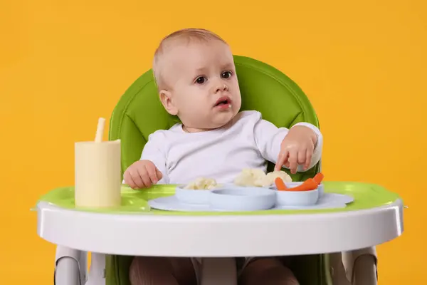 Cute little baby with healthy food in high chair on orange background