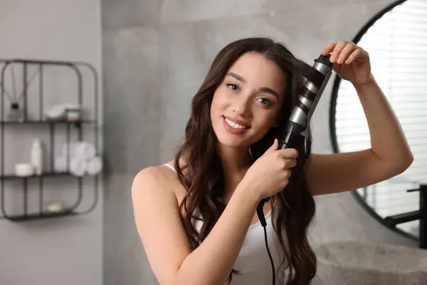 Smiling woman using curling hair iron in bathroom