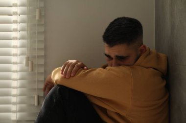 Evdeki pencerenin yanında oturan üzgün adam.