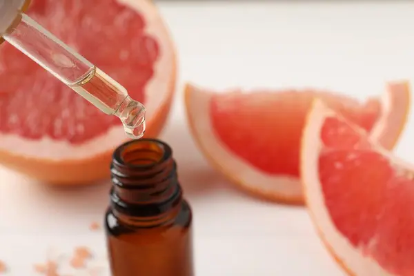 Dripping grapefruit essential oil from pipette into bottle and fresh fruit on white table, closeup. Space for text
