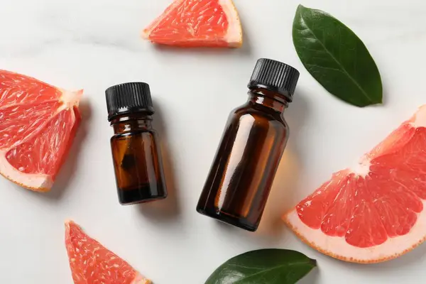 Grapefruit essential oil in bottles, leaves and cut fruit on white marble table, flat lay