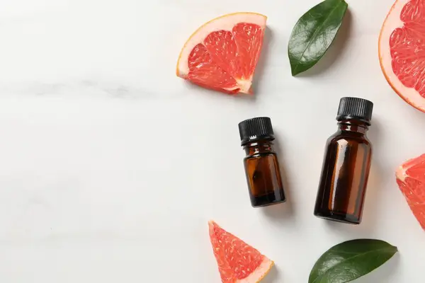 Grapefruit essential oil in bottles, leaves and cut fruit on white marble table, flat lay. Space for text