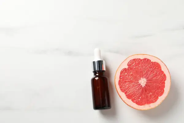 Grapefruit essential oil in bottle and half of fruit on white marble table, top view. Space for text