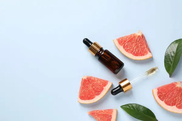 Grapefruit essential oil in bottle, pipette, leaves and cut fruit on light blue table, flat lay. Space for text