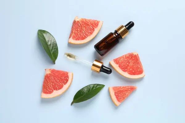 Grapefruit essential oil in bottle, pipette, leaves and cut fruit on light blue table, flat lay