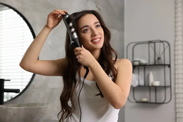Smiling woman using curling hair iron in bathroom
