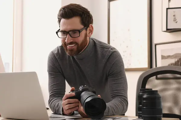 Professional photographer holding digital camera near table with laptop indoors
