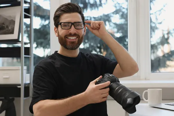 Professional photographer in glasses holding digital camera at table indoors
