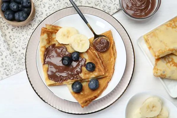 Tasty crepes with chocolate paste, blueberries and banana served on white wooden table, flat lay