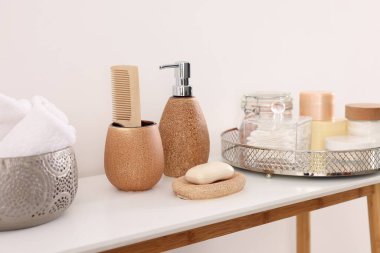 Beyaz duvarın yanındaki masada farklı banyo aksesuarları ve kişisel bakım ürünleri var.