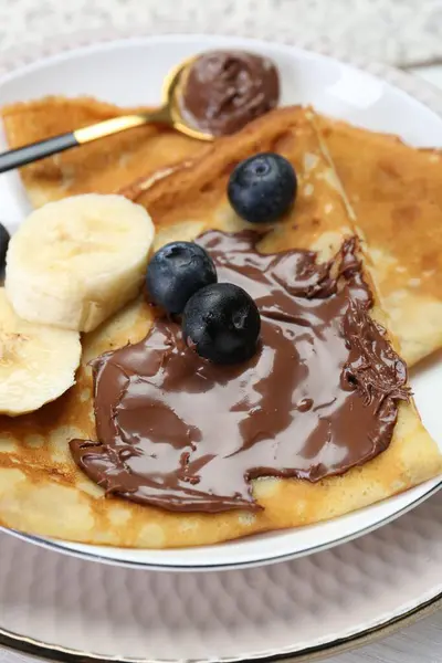 Tasty crepes with chocolate paste, blueberries and banana served on white wooden table, closeup