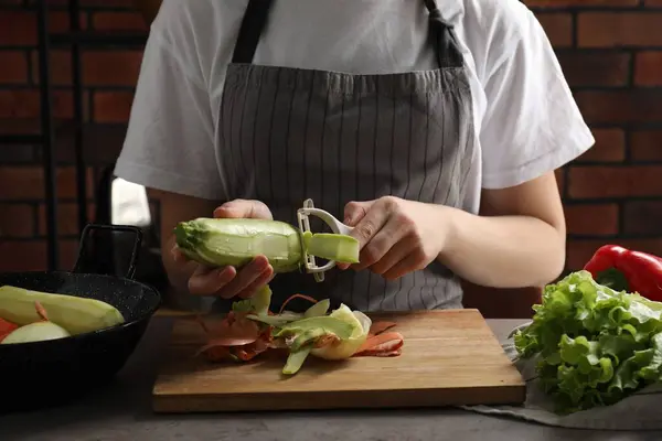 Woman peeling fresh zucchini at grey table indoors, closeup