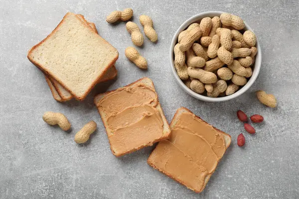 Tasty peanut butter sandwiches and peanuts on gray table, flat lay