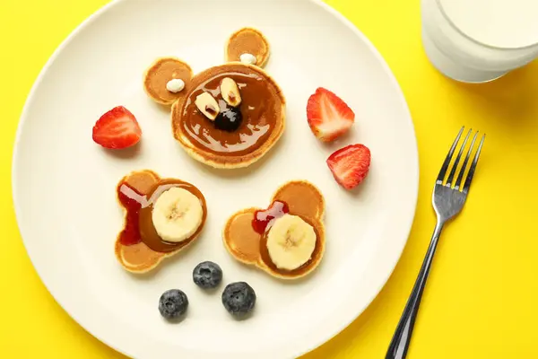Creative serving for kids. Plate with cute bears made of pancakes, berries, banana and chocolate paste on yellow background, flat lay