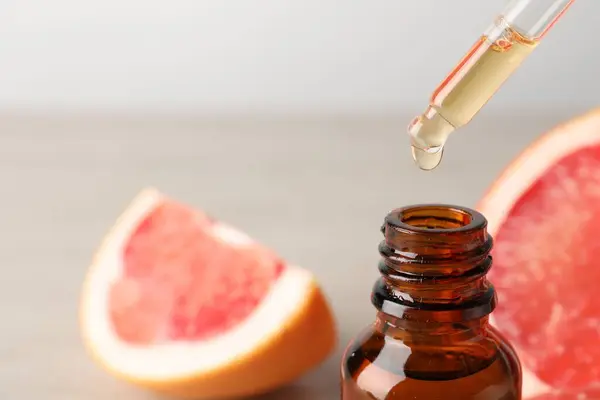 Dripping grapefruit essential oil from pipette into bottle and fresh fruit on table, closeup. Space for text