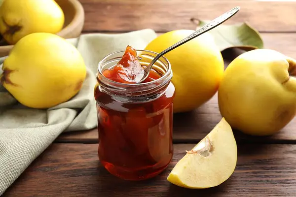 Tasty homemade quince jam in jar, spoon and fruits on wooden table