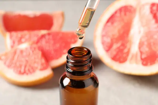 Dripping grapefruit essential oil from pipette into bottle and fresh fruit on light table, closeup