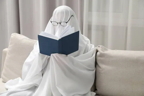 Creepy ghost. Person covered with white sheet reading book on sofa at home