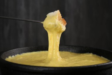 Bir parça ekmeği fondü tenceresine daldırırken lezzetli eritilmiş peynirle koyu gri arka planda, yakın plan