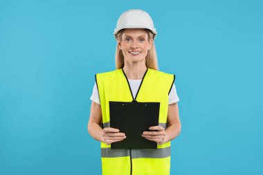 Açık mavi arka planda elinde pano tutan sabit şapkalı mühendis.