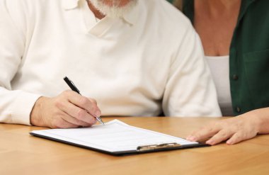 Ahşap masada son dilek ve vasiyetnameyi imzalayan yaşlı çift, yakın plan.