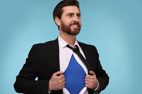 Happy businessman wearing superhero costume under suit on light blue background
