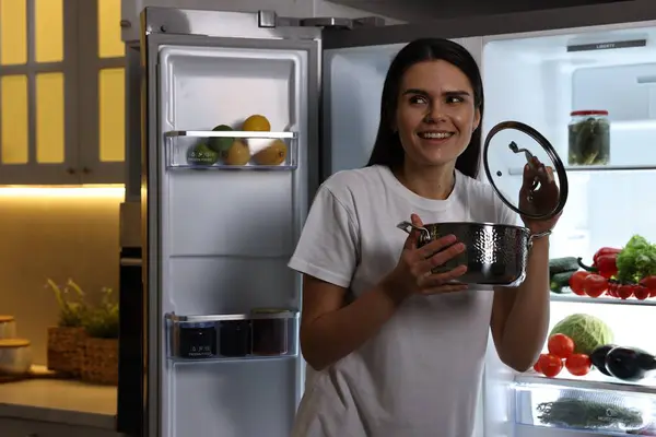 Young woman with pot near modern refrigerator in kitchen at night
