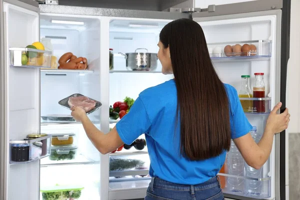 Young woman taking pack of meat out of refrigerator indoors, back view