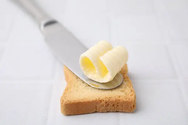 Tasty butter curl, knife and piece of dry bread on white tiled table, closeup