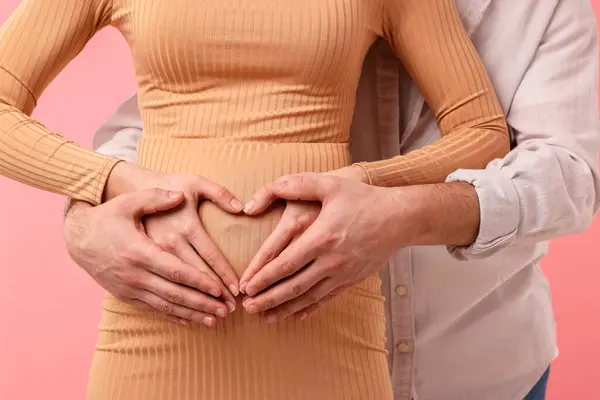 妊娠中の女性と彼女の夫は ピンクの背景に対する腹の手で心を作ります クローズアップ — ストック写真