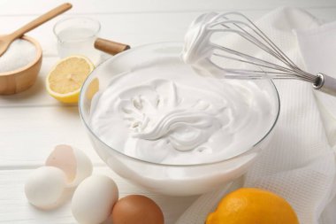 Krem şantili kase, çırpma ve malzemeler beyaz ahşap masada.