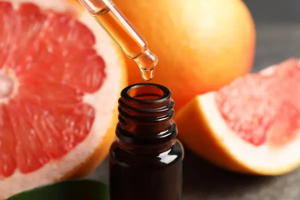 Dripping grapefruit essential oil from pipette into bottle and fresh fruits on table, closeup