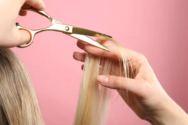 Hairdresser cutting client\'s hair with scissors on pink background, closeup