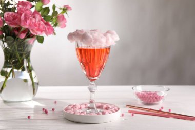 Cam kaplı pamuk şeker kokteyli, marşmelov, pembe güllü vazo ve gri arka planda beyaz ahşap masa üzerinde pipet.