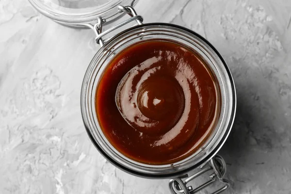 Tasty barbeque sauce in open jar on grey textured table, top view