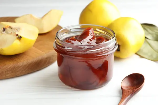 Tasty homemade quince jam in jar, spoon and fruits on white wooden table, closeup