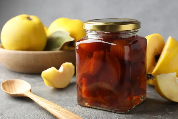 Tasty homemade quince jam in jar, spoon and fruits on grey textured table, closeup