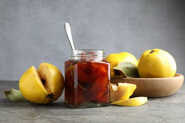Tasty homemade quince jam in jar, spoon and fruits on grey textured table