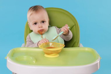 Küçük şirin bebek, açık mavi arka planda, yüksek sandalyede sağlıklı yiyecekler yiyor.