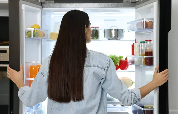 Young woman near modern refrigerator in kitchen, back view