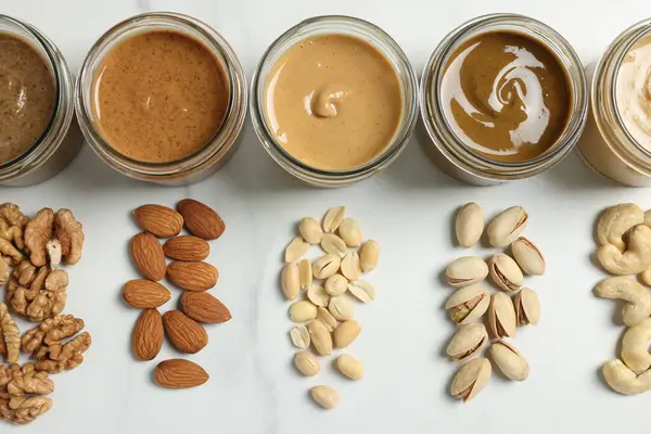 Kavanozlarda lezzetli fındık ezmesi ve beyaz mermer masada çiğ fındık, düz uzanmış