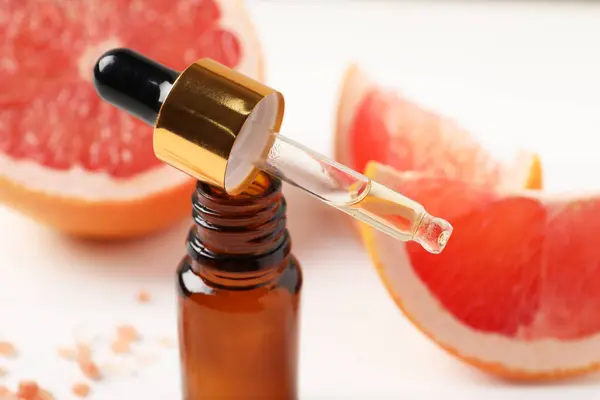 Grapefruit essential oil in bottle, pipette and fruit on white table, closeup