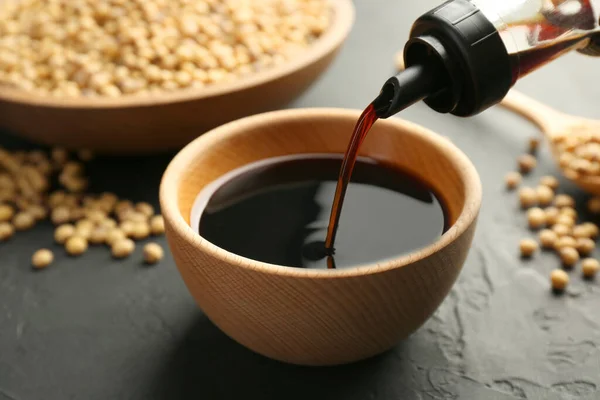 Pouring tasty soy sauce from bottle into bowl on black table, closeup